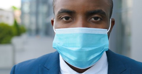 Close up of face of young African American handsome businessman in medical mask looking straight to camera with happy look. Portrait of man at street. Coronavirus concept.