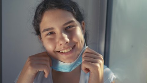 coronavirus pandemic concept. little teenage girl kid takes off medical mask and smiles a beautiful sunlight from the window. self-isolation virus covid 19 infection doomsday. coronavirus child 