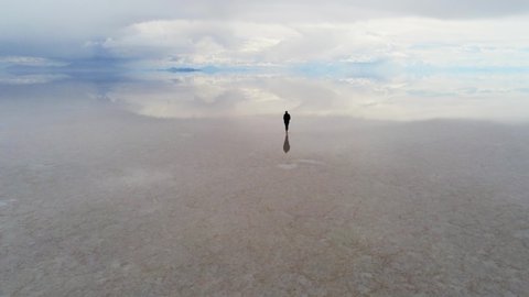 Aerial View of Female Silhouette Walking on Wet Uyuni Salt Flat, Bolivia. Endless Horizon and Mirror Reflection of Sky in Background