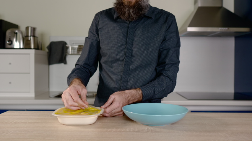 torso of a man with a beard opening a container of ready made food and throwing it on a plate
