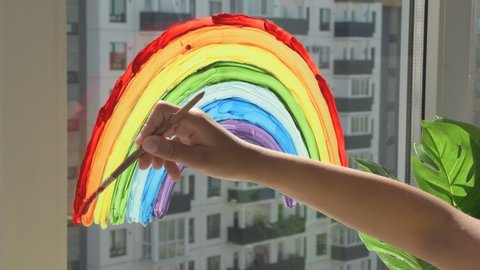 Girl painting rainbow on window during quarantine at home.