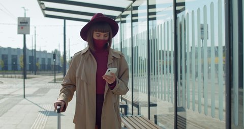 Stylish girl in protective mask and suitcase using smartphone near airport. Millennial female passenger walking and looking at phone screen. Concept of coronavirus and travelling