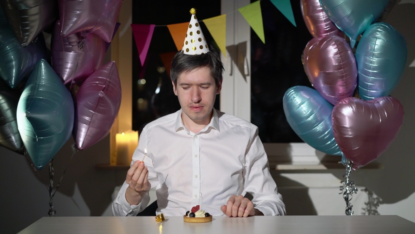 Young man celebrating birthday alone. Sad bored and tired male bday with party cake and blowing candle, balloons