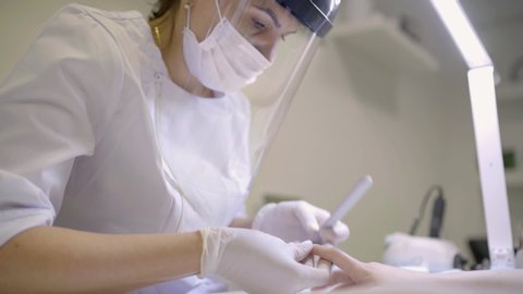 Professional manicure master in Transparent Safety Face Shield and mask using nail file grinding down nails during manicure procedure. Small business existence at COVID-19 lockdown concept video. 