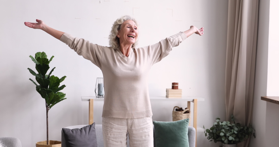 Excited middle aged 70s beautiful healthy woman standing with outstretched arms, feeling emotional indoors. Happy elderly mature grandmother welcoming new day, doing morning stretching exercises. Royalty-Free Stock Footage #1053479321