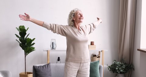 Excited middle aged 70s beautiful healthy woman standing with outstretched arms, feeling emotional indoors. Happy elderly mature grandmother welcoming new day, doing morning stretching exercises.