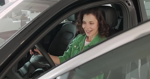 Young beautiful girl chooses new car at car dealership. The woman is happy, smiling and rejoicing in the purchase new car. Girl in awe of new vehicle. The buyer selects a car in the catalog.
