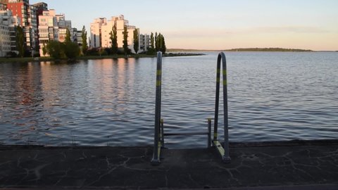 Focus on pier ladder and calm water in the lake. In the background is the sunset light coming over residential buildings located next to lake Malaren in Vasteras city, Sweden