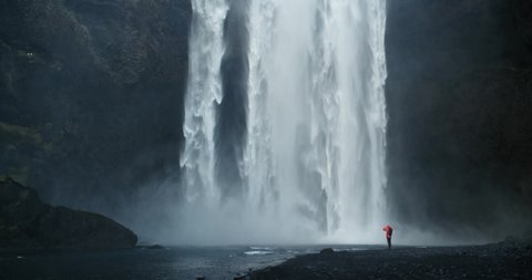Man standing under majestic Skogafoss waterfall. Huge amount of water falling down from high cliff. Golden Circle attraction. Traveling in Iceland during holiday trip. Handheld shot in slow-motion