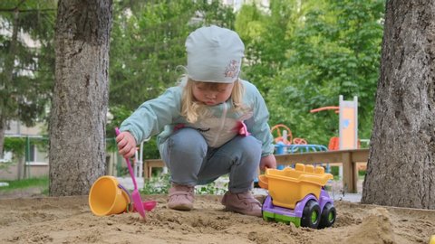 Cute little blond girl playing alone with plastic toy car and sand tools in sandbox
