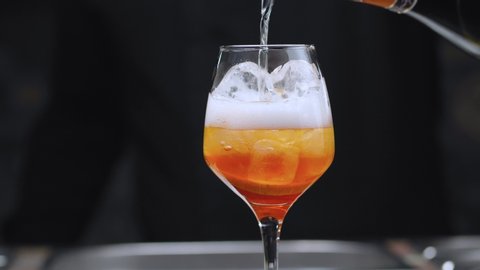 Cooking an Aperol Spritz Cocktail. Close up bartenders hands pouring prosecco in wine glass with ice, mixing cocktail with the spoon sprinkling it with orange essential oil. Long fizzy drink.