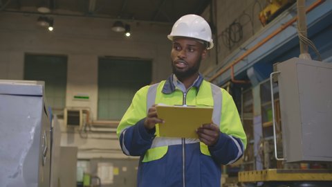 Portrait of afro-american supervisor writing down report in clipboard during inspection of manufacturing plant at industrial factory. African engineer doing inventory