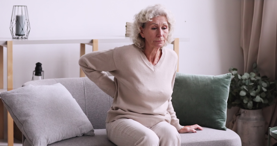 Unhappy middle aged 70s grey haired woman feeling lower lumbago spinal backpain while getting up from couch. Unhealthy senior mature retired grandmother suffering from osteoporosis arthritis. | Shutterstock HD Video #1053499907