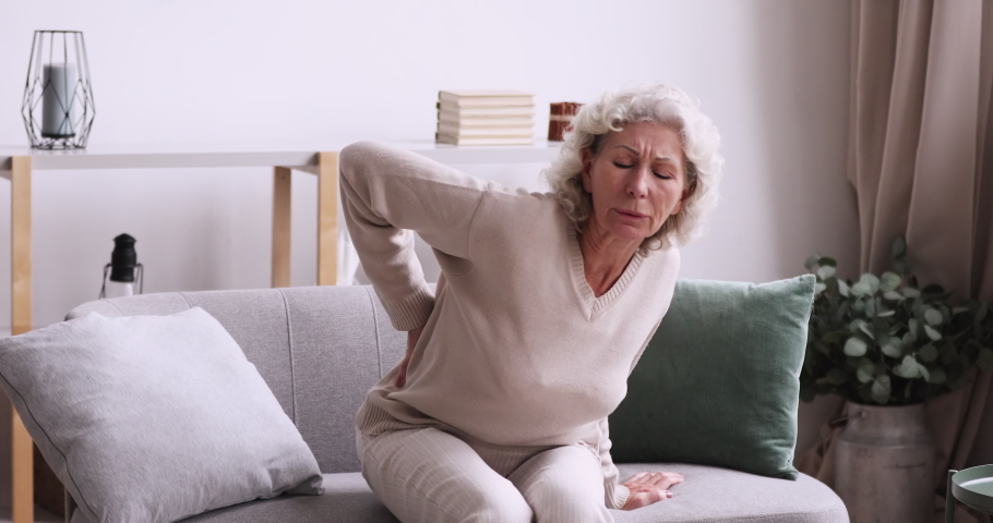 Unhappy middle aged 70s grey haired woman feeling lower lumbago spinal backpain while getting up from couch. Unhealthy senior mature retired grandmother suffering from osteoporosis arthritis.