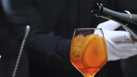 Preparing an Aperol Spritz Cocktail. Close up bartenders hands pouring prosecco in wine glass with ice and sprinkling cocktail with orange essential oil. Long fizzy drink.