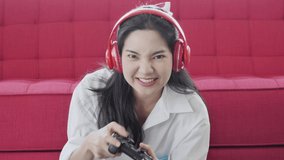 Young Asian woman with a smiling face wearing a white shirt sitting in front of the red sofa in the living room, she is excited about playing computer games.