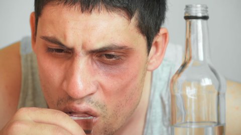 Drunkard young guy with bruised eye drinks an alcoholic drink at the table at home. The problem of alcohol addiction. Withdrawal pains. Close up. portrait.