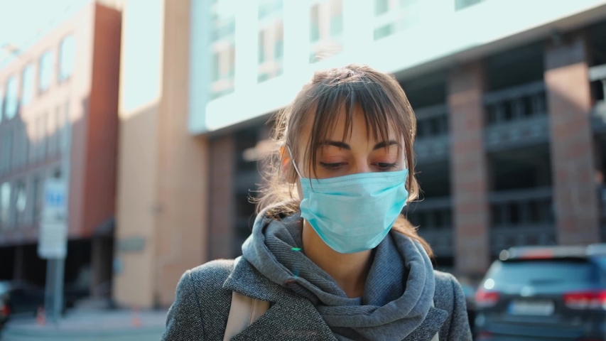 slow motion mixed race brunette girl in grey coat and protective face mask walks outdoors on city street sunny day with buildings on background. still quarantine Royalty-Free Stock Footage #1053502658