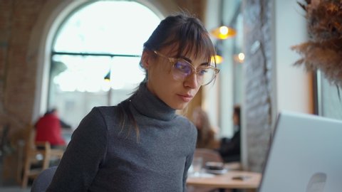Portrait young woman in eyeglasses using laptop in coworking space or cafe and drinking coffee. female sitting in front open laptop computer. Study, learning, remote work, freelance.
