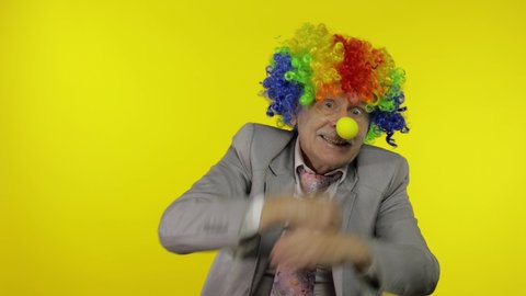 Senior clown manager office worker in wig and business suit at work dancing, entertains, celebrate victory, making silly faces. Guy businessman freelancer boss. Halloween. Yellow background