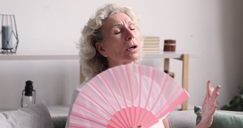 Head shot anxious mature elderly woman using paper fan, feeling overheated at home. Nervous middle aged senior lady cooling herself, suffering from hot summer weather indoors without air conditioner.