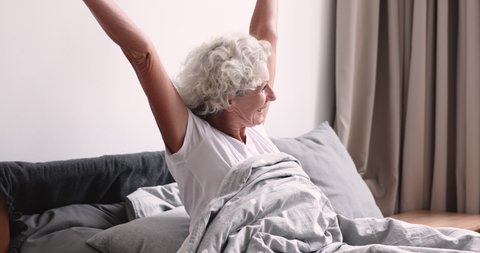 Happy refreshed middle aged woman waking up in morning, stretching back sitting in bed, welcoming new day alone at home. Smiling mature elderly lady feeling energetic after good night sleep rest.