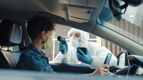 Man in medical suit is taking body temperature of woman driver in car outdoors during covid-19 pandemic, guy is using no-touch infrared thermometer.