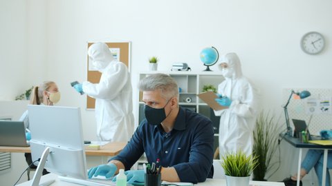People in protective suits are taking body temperature in office with infrared thermometer then finding infected guy in mask asking him to follow with testing and quarantine.