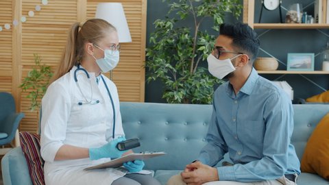 Female doctor young blond woman in uniform is taking male patient's temperature with infrared thermometer in apartment, people are wearing masks.