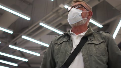 Portrait of a senior citizen in a mask, glasses against the ceiling with fluorescent lamps. Post-quarantine life, social distance. Portrait of old male wearing face mask during coronavirus outbreak