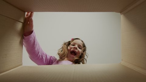 Little Girl Opens Cardboard Box And Shouts Happily. She Is Surprised. View From Inside The Box.