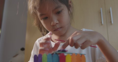 Asian little girl glueing colored ice cream sticks by hot melt electrical glue gun, close up, slow motion moving camera. Elementary school crafts class. Children have fun on a handicraft project.