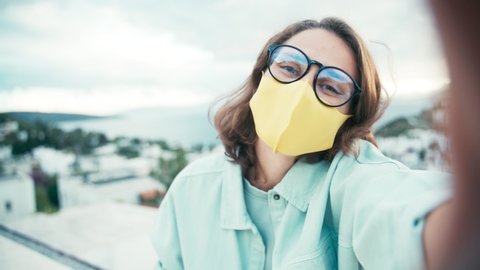 Young cheerful woman in glasses and bright yellow face mask recording video for vlog in social media or making a video call.