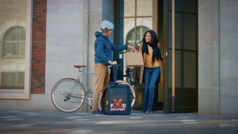 Happy Food Delivery Man Wearing Thermal Backpack Rides Bike to Deliver Restaurant Order to a Beautiful Female Customer. Courier Delivers Lunch to Gorgeous Girl in Modern City District Office Building