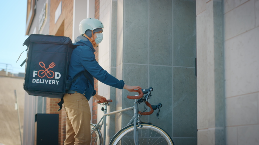 Food Delivery Man Wearing Protective Medical Face Mask and Thermal Backpack Rides a Bike to Deliver Order to a Masked Female Customer. Courier Delivers Takeaway Lunch. Quarantine, Social Distancing | Shutterstock HD Video #1053526517