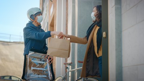 Food Delivery Man Wearing Protective Medical Face Mask and Thermal Backpack Rides a Bike to Deliver Order to a Masked Female Customer. Courier Delivers Takeaway Lunch. Quarantine, Social Distancing