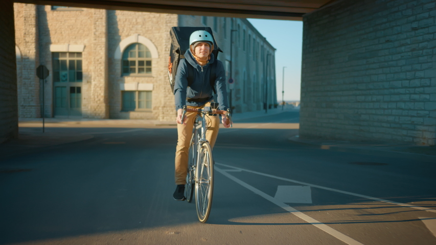 Happy Food Delivery Courier Wearing Thermal Backpack Rides a Bike on the Road To Deliver Orders and Packages for Clients. Sunny Day in Modern City with Stylish Urban Buildings. Front Following Shot Royalty-Free Stock Footage #1053526547