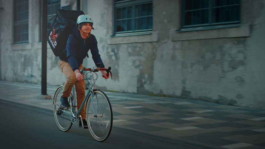 Happy Food Delivery Courier Wearing Thermal Backpack Rides a Bike on the Road To Deliver Orders for Clients and Customers. Sunny Day in Modern City with Stylish Urban Buildings. Front Following Shot | Shutterstock HD Video #1053526550