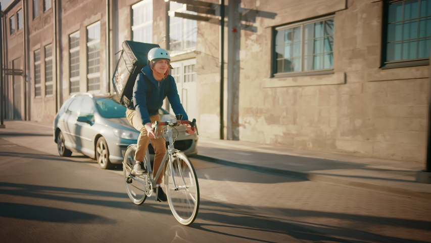 Happy Food Delivery Courier Wearing Thermal Backpack Rides a Bike on the Road To Deliver Orders for Clients and Customers. Sunny Day in Modern City District with Buildings. Slow Motion Front Followig Royalty-Free Stock Footage #1053526553