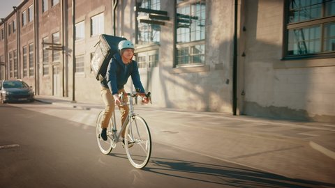Happy Food Delivery Courier Wearing Thermal Backpack Rides a Bike on the Road To Deliver Orders for Clients and Customers. Sunny Day in Modern City District with Buildings. Slow Motion Front Followig