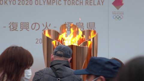 FUKUSHIMA, JAPAN - 24 MAR 2020 : Olympic Flame displayed at Fukushima station. Tokyo Olympic 2020 have been postponed to 2021 due to coronavirus. Crowd of people wearing surgical masks. Slow motion.