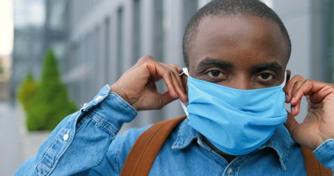 Portrait of young African American handsome and cheerful man taking on medical mask and looking at camera. Pandemic of coronavirus concept. Male putting on respiratory protection in city.