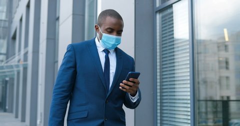 African American businessman in medical mask walking the street and texting message on mobile phone. Male pedestrian in respiratory protection strolling outdoor and tapping or scrolling on smartphone.