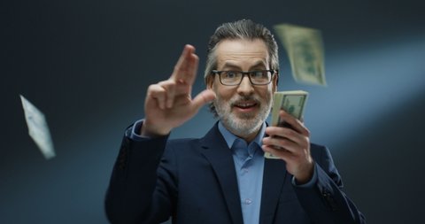 Portrait shot of cheerful rich handsome businessman of middle-age in glasses and suit tossing money on grey wall background. Happy good looking man in business style throwing dollars.