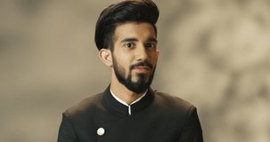 Close up of young happy cheerful handsome Hindu man with beard and in black jacket looking at camera and smiling joyfully. Portrait of Indian attractive joyful male with smile.