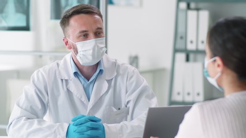 Male doctor in protective face mask, disposable gloves and lab coat speaking with female patient during medical consultation in clinic. Coronavirus pandemic concept