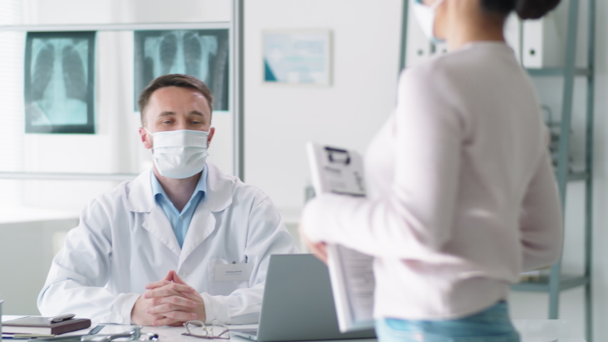 Male doctor in protective mask and lab coat sitting at desk in clinic, reading medical history of female patient and speaking with her during consultation. Covid-19 pandemic concept Royalty-Free Stock Footage #1053539963