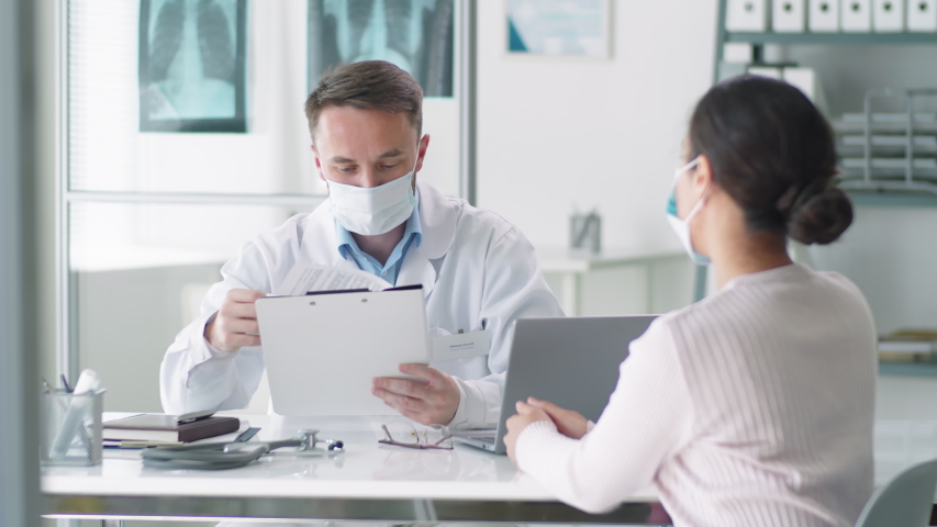 Male doctor in protective mask and lab coat sitting at desk in clinic, reading medical history of female patient and speaking with her during consultation. Covid-19 pandemic concept | Shutterstock HD Video #1053539963