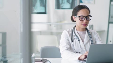 Attractive mixed-raced female doctor in lab coat, glasses and stethoscope over her neck using laptop at desk, looking at camera and smiling while working in medical office