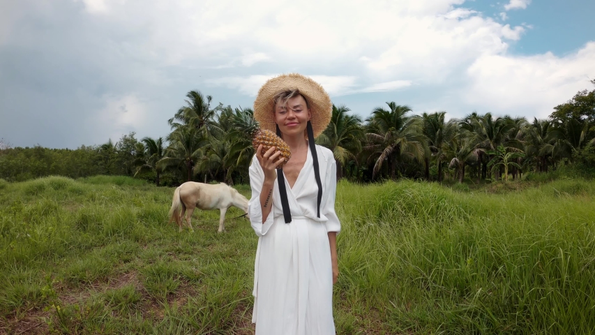 Girl with a pineapple in hand, in a white dress and a straw hat stands in a field next to the horse